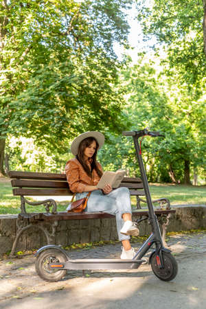 A young beautiful girl with a hat is sitting on a park bench reading a book. An electric scooter is parked next to it, while nature and green colors predominate in the background.の写真素材