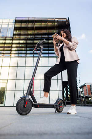 A young beautiful girl is standing next to her electric scooter while typing on her phone. She is smiling, business-trained, with modern architecture in the background.の写真素材