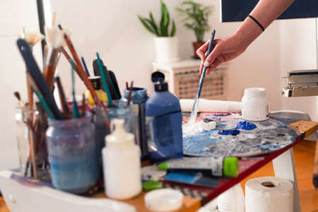 The painter's hand smears paint on a palette in her home studio. The paint pots and the painting brush are in jars on the side.の写真素材