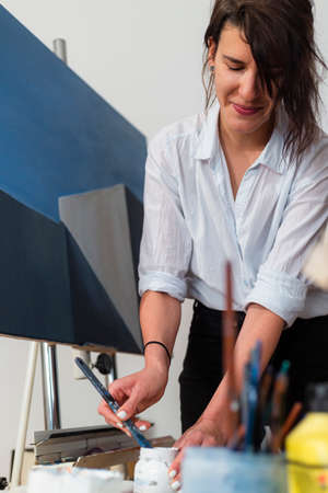 A young painter squeezes paint on a palette in her home studio. Behind it is a work of art placed on an easel, as well as other accompanying material for painting.の写真素材