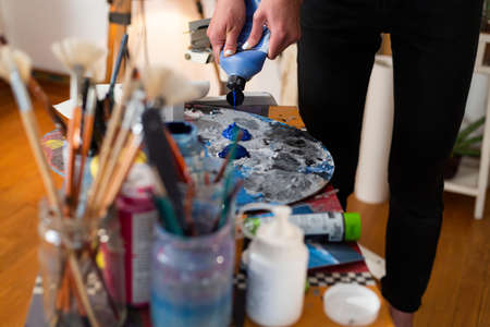 Close up of painter's hand extruding color on a palette in her studio. Around the palette are a multitude of brushes for painting in jars.の写真素材