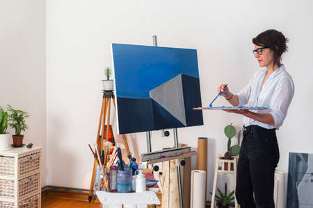 A young smiling painter mixes colors on a palette in her home studio. Behind it, you can see a contemporary work of art placed on an easel, as well as other accompanying material for painting.の写真素材
