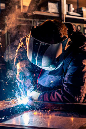 Close up of a welder welding in workshop. He is dressed in a work uniform and has a protective...