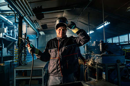 Portrait of a smiling welder posing in workshop while his colleague in the background is working with grinder. They are both in work uniforms and have protective helmets on their heads.の写真素材