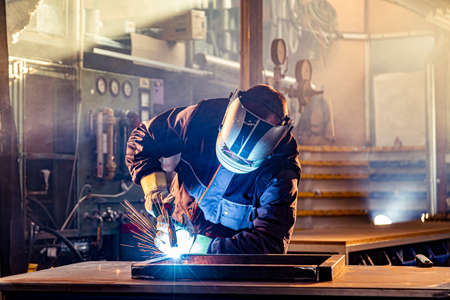 The welder performs welding task at his workplace in the factory, while the sparks fly around, he...