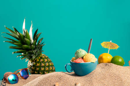 A cup with a few scoops of ice cream in the sand. In addition, you can also see fruits such as pineapple, lemon, lime, but also sunglasses and seashells. The background is also half in the sand, half in the blue wall/sky.の写真素材