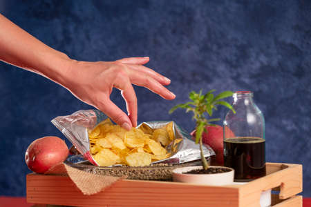 A hand picking up chips from an open package, CBD oil in a bottle, potatoes, a marijuana plant and a bowl of hemp seeds in a wooden crate. The background is blue.の写真素材
