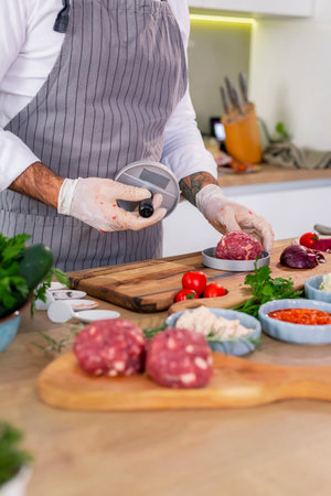 A close-up of a chef shaping meat for burgers in a press on a wooden board in the kitchen. There are various spices, vegetables, as well as small kitchen bowls around the work surface.の写真素材