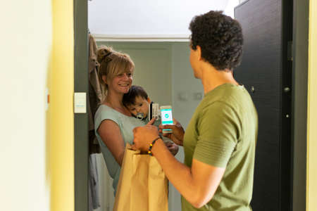 A mother with her baby in her arms pays with a card to a delivery person via a smart phone at the...