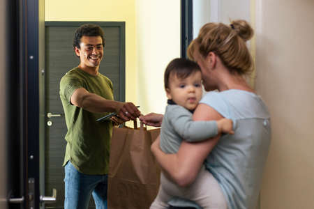A mother with a baby in her arms at the door of her home picks up the package delivered by a...