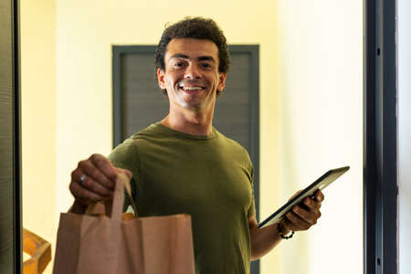 A smiling delivery man holds a tablet in his hand and hands out a paper bag at the entrance to the apartment in the building.の写真素材