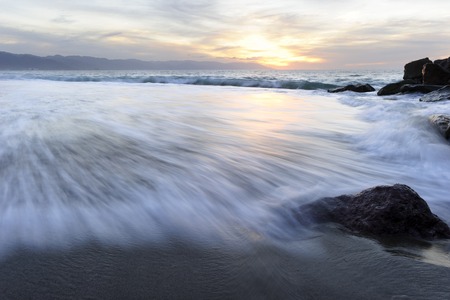 Ocean Sunset beach waves is an images of a rush of waves captured by a time exposure with a glowing orangesunset cloud filled sky on the ocean horizon.の写真素材