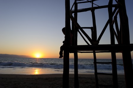Boy childhood silhouette is boy sitting in his place of of thinking, dreaming and reflection at the beach as the sun sets on the ocean horizon.の写真素材