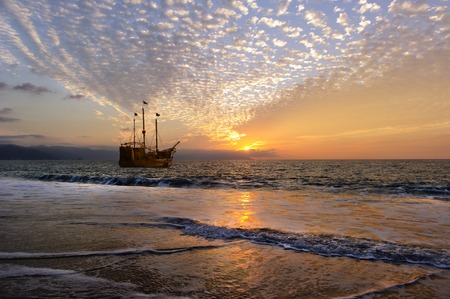 Pirate Ship is an old wooden pirate ship with full flags as the sun sets on the ocean horizon in a colorful sunset sky.