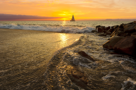 Sailboat sunset is a silhoueette of a boat sailing with a colorful sunset sky and the ocean waves breaking on the sea shore.の写真素材