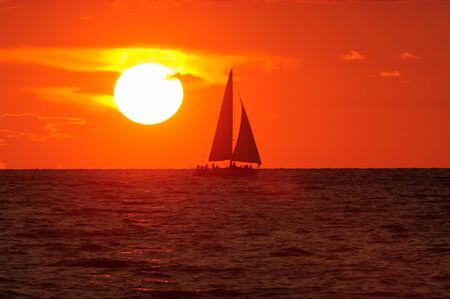 A Sailboat is sailing along the water silhouetted against an orange sunset sky in a peaceful serene nature landscape.の写真素材