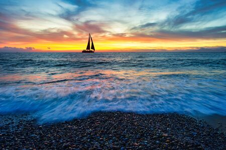 A Sailboat is Sailing Along the Ocean as a Vivid Colorful Sunset is in the Backgroundの写真素材