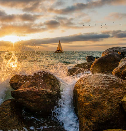 A Sailboat Is Sailing Along The Ocean Sunset As A Wave Splashes On Shore In A Vertical Image Formatの写真素材