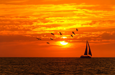 Colorful Image Of A Sailboat Sailing Along The Ocean Horizon With Orange Sunset Skyの写真素材