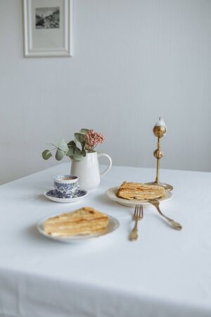 two pieces of cake on white plates golden forks a vase with a flower and coffee and a golden candlestick on a white tableの写真素材