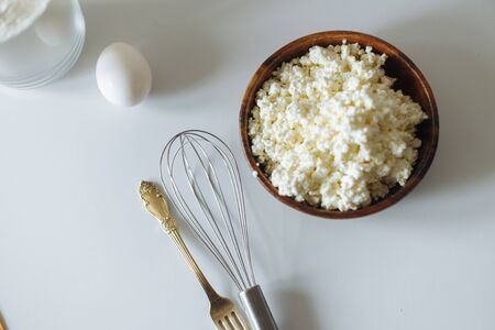 cottage cheese in a wooden cup golden fork and spoon .kitchen whisk and egg on a white tableの写真素材