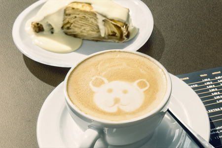 Cappuccino with little bear decoration drawn into the foam. In the background the austrian specialty \\\"Apfelstrudel\\\" with vanilla sauce.の写真素材