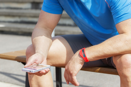 Sportive man sits on a bench and checks his fitness results on a smartphone. He wears a fitness...