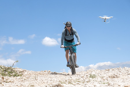 Mountain biker with helmet camera rides a bright gravel road and is followed by a white droneの写真素材