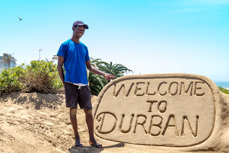 DURBAN, SOUTH AFRICA- NOVEMBER 02, 2017: Unknown sand sculpture builder at bay Plenty in Durban stands beside his artwork and points to it.のeditorial素材