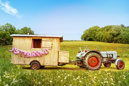 Scenic placed wooden trailer with tractor and pink "just married" text balloons on flower covered meadow anouncing happy marriage.の写真素材