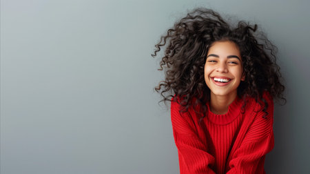 A joyous young woman wearing a vivid red sweater, laughing with curly hair flowing on a gray background, showing happiness and warmth.の素材