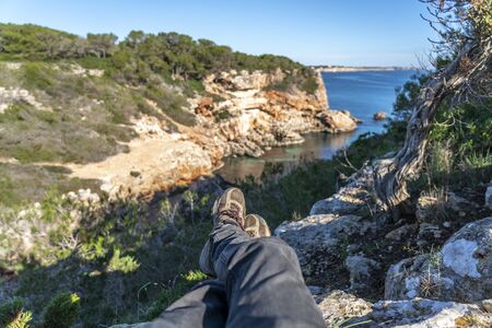 Pov view of two male legs with mountain boots of a man lying resting on a cliff of a cove of the Mondrago natural park, in Mallorca. Unfocused mediterranean typical landscape in the background.の写真素材