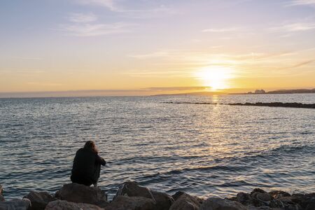 Sea landscape from a beach in the promenada of Palma city, Majorca. A girl sitting on the rocks on her back, watching the sunset in meditation poseの写真素材