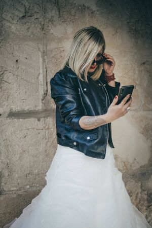 Blonde mature beautiful woman wearing a wedding dress, a leather jacket, looking at a notification on her smartphone with indignant countenance while holding her sunglasses. Rocker look.の写真素材