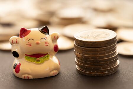 Maneki neko or beckoning with a coin stack, and more unfocused coins on the background.. Conceptual symbol of luck, abundance and economic success.の写真素材