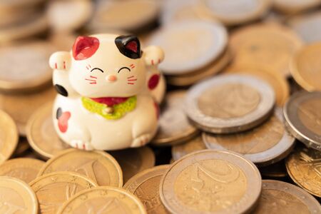Maneki neko or beckoning cat placed on a pile of scattered coins. Conceptual symbol of luck,...