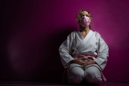 Woman with mask in karate pose sitting on her knees in gym hall, pink background.
