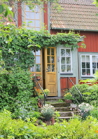 Wooden red cottage entrance with many windows and door ajar in the countryside of Sweden, surrounded by beautiful greenery at summer time.の写真素材
