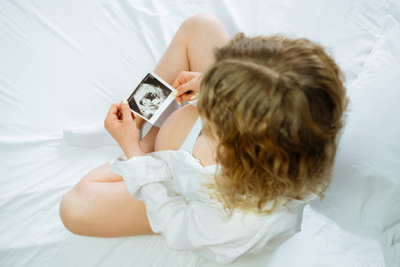 The pregnant girl holding an ultrasound picture in her hands while sitting on the bedの写真素材