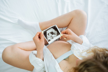 The pregnant girl holding an ultrasound picture in her hands while sitting on the bedの写真素材