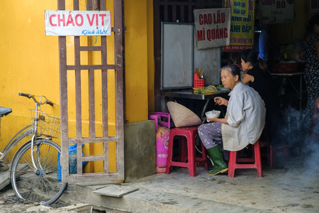 Hoi An / Vietnam, 11/11/2017: Local Vietnamese woman with rice hat and bicycle eating in a street food stall in Hoi An in Vietnam.のeditorial素材