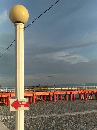 Toilet shower is written on a sign with an arrow indicating the direction of the shower and toilet. Announcement on a white pole against a red metal pier and a wooden beach path. boats boats in the background and the sea ocean, Golden hour, grey skyの写真素材