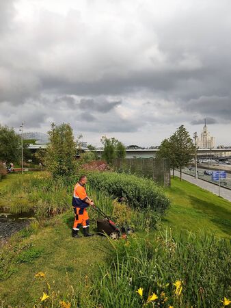 Moscow, Russia - July 25, 2019: text Gastronomic center. Zaryadye Park in Moscow. A man in uniform and rubber boots mows the lawn with a lawnmower. In the foreground green bushes, trees, grass, colorful flowers. Distant plan high-rise building with a spirのeditorial素材