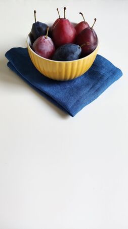 A yellow ceramic bowl with red and blue ripe plums on a white table against a washed-out gray wall. The plate is on a blue linen napkin. Side view, food photography.の写真素材