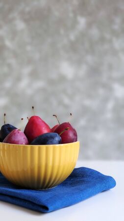 A yellow ceramic bowl with red and blue ripe plums on a white table against a gray wall. The plate is on a blue linen napkin. front view, food photography. Items in the lower left corner.の写真素材
