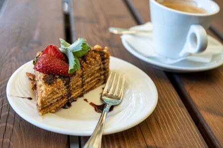 a cup of black coffee and a piece of honey sponge cake decorated with strawberries and mint on a wooden tableの写真素材