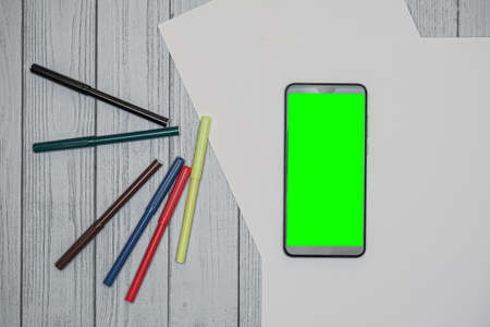 cell phone with green screen with felt-tip pens and white sheets on a wooden table. Mobile app...