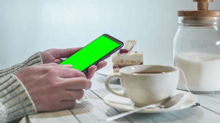Woman holding smartphone with green screen with hot tea and cake on table