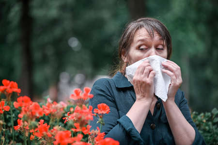 A woman blows her nose in a handkerchief in the garden with flowers in the foreground. Spring allergy concept.の写真素材