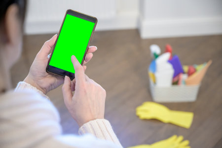 a woman holds a phone against the background of a plastic basket with detergents, brushes, sponges and rubber gloves for cleaning.の写真素材